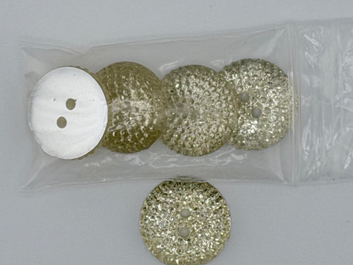 Three round, glittery gold buttons with a white button, all inside a clear plastic bag, with one loose gold button placed to the left on a light gray background.