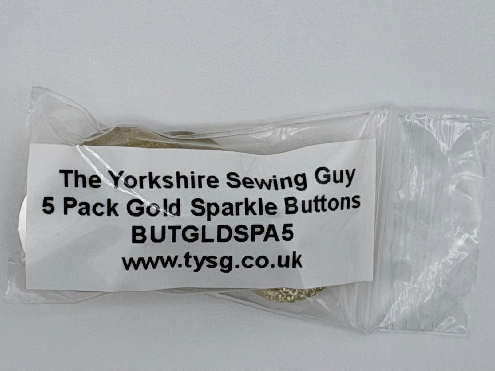 A clear plastic bag containing five small gold buttons, with a white paper label printed with the text 'The Yorkshire Sewing Guy 5 Pack Gold Sparkle Buttons BUTGLDPS5 www.tysg.co.uk