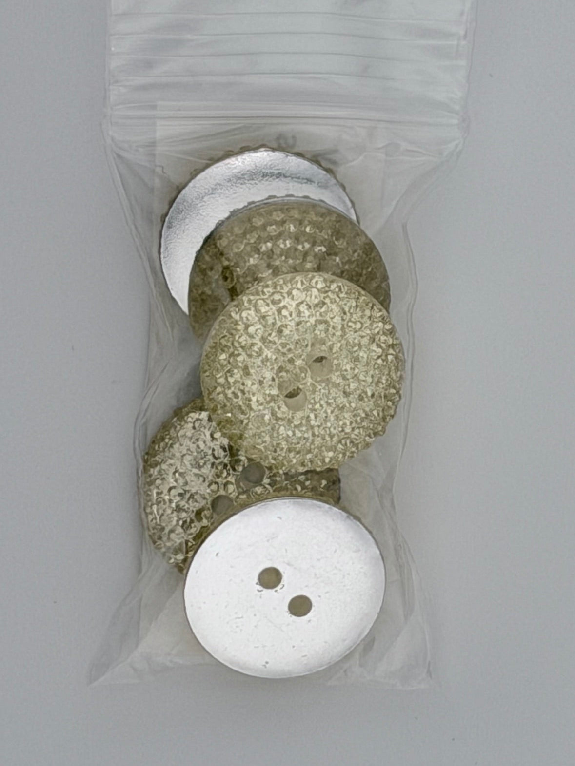 A small clear plastic bag containing several decorative buttons on a plain light gray background. The buttons include textured metallic silver, textured champagne-gold, and a solid white button with two holes.