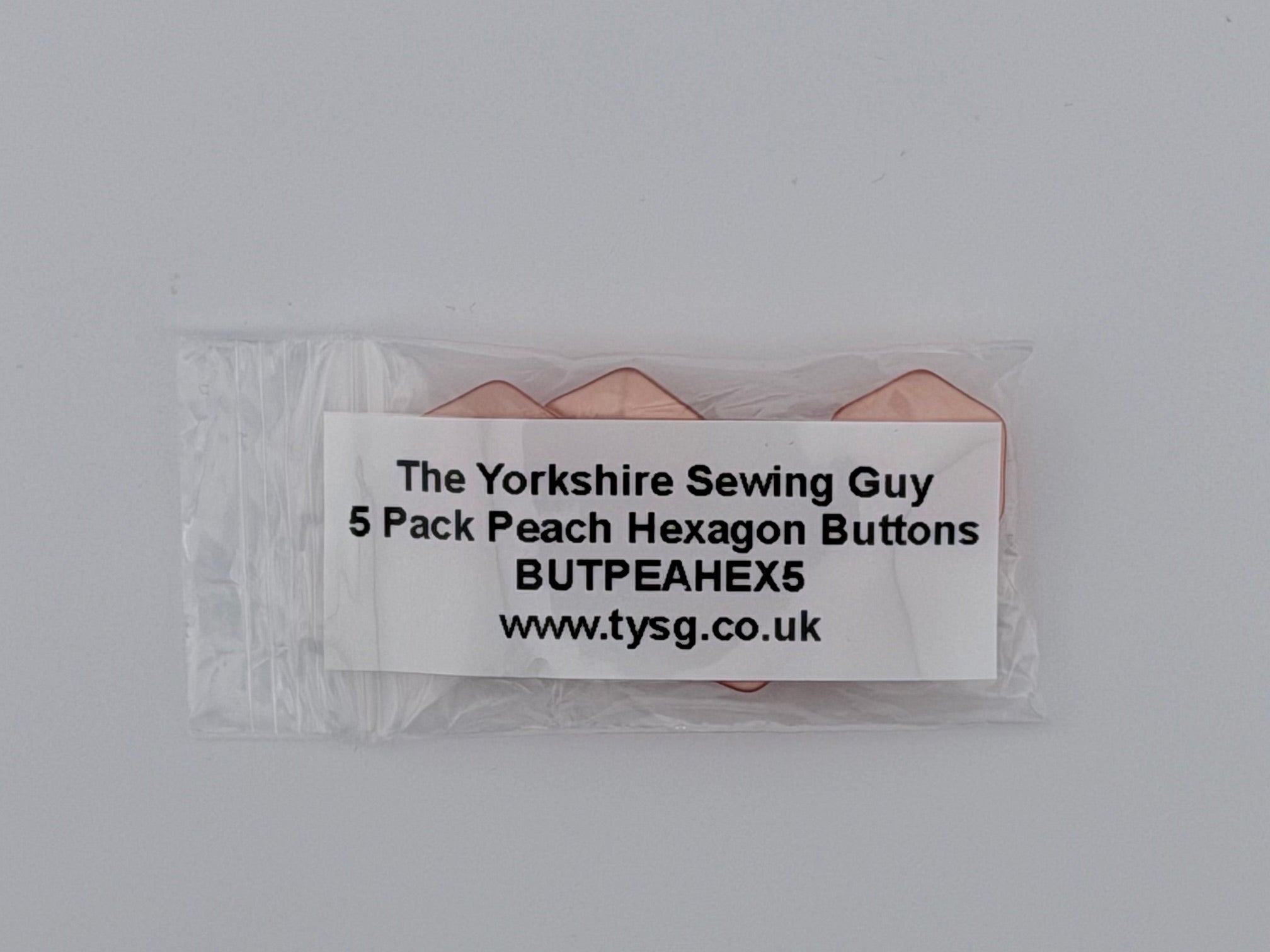A small clear plastic bag containing five peach-colored hexagon buttons, with a white paper label that reads 'The Yorkshire Sewing Guy 5 Pack Peach Hexagon Buttons BUTPEAHEXS www.tysg.